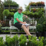 Man sitting on a bench at a garden center with plants around, listening to customer stories.