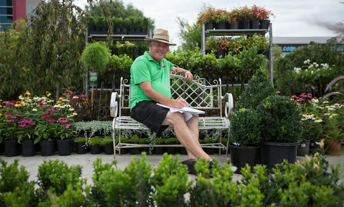 Man sitting on a bench at a garden center with plants around, listening to customer stories.