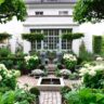 A well-manicured garden with hydrangeas and topiaries in front of a white house, featuring a small water fountain and a symmetric brick pathway, ideal for garden makeovers.