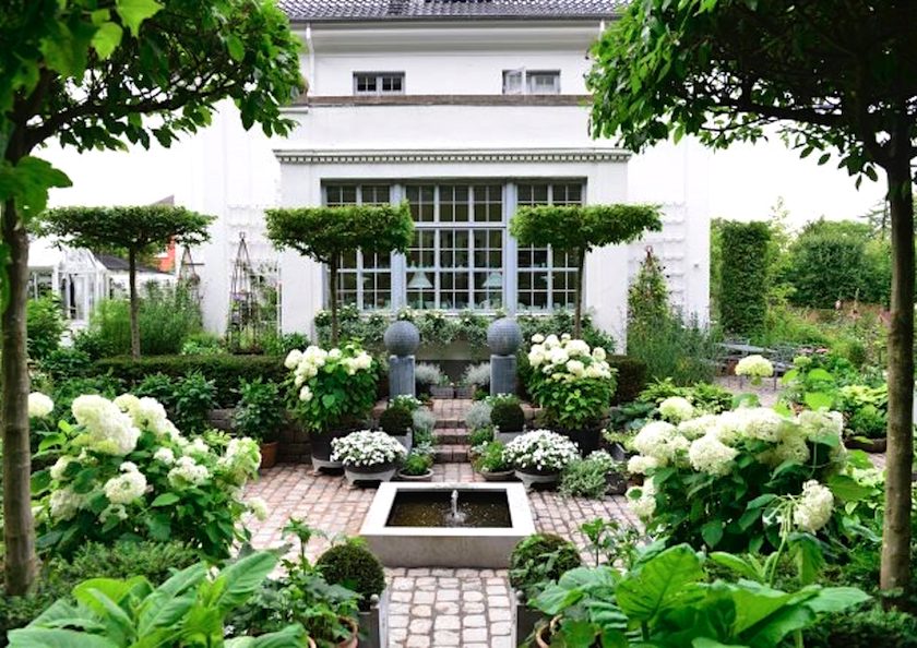 A well-manicured garden with hydrangeas and topiaries in front of a white house, featuring a small water fountain and a symmetric brick pathway, ideal for garden makeovers.