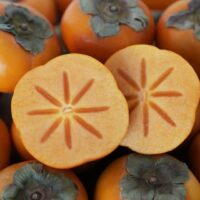 Close-up of ripe Persimmon 'Fuyu' 7L Bag with a sliced one in the foreground showing the star-shaped pattern inside.