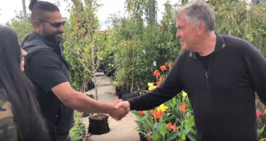Two men shaking hands in a garden design center, surrounded by plants and flowers, with a woman partially visible on the left.