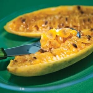 Close-up of a halved Passionfruit Banana Vine 6" Pot on a green plate with a spoon, showcasing its juicy seeds and vibrant yellow pulp from a passionfruit vine.