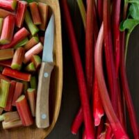 Chopped rhubarb pieces and a knife in a wooden bowl next to whole Rhubarb stalks on a dark surface, including the lush Rhubarb 'Glaskins Perpetual' 6" Pot.