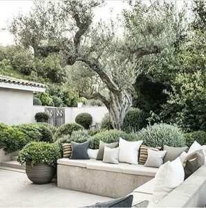 An elegant outdoor seating area with plush cushions on a built-in stone bench, surrounded by a lush planting garden and trees.