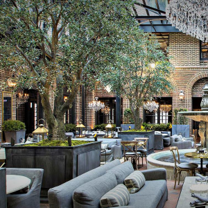 Elegant courtyard dining area with a planting garden, plush sofas, and hanging lights, framed by brick walls and arched windows.