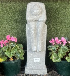 A white stone sculpture of a figure with its head resting on its knees is placed between two pots of pink flowers against a green background, reminiscent of an Iridami Resting Man Grey Terrazzo 16x20x60cm.