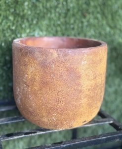 A large, round Portica Egg Pot Rust M 16x13cm with a rough texture sits on a black metal stand against a background of green artificial grass, making it an ideal garden planter.