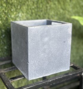 A plain, gray, square concrete planter sits on a metal rack with a backdrop of green foliage, reminiscent of the sleek design of the Portica Egg Pot Rust M 16x13cm (Copy).