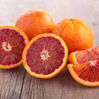 Two whole and two halved blood oranges, displaying vibrant red flesh, rest on a wooden surface. These fruits highlight the rich hue and succulent nature of the prized Citrus Orange Tree 'Blood Orange' Dwarf from a 10" pot.