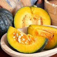 A cut Pumpkin 'Japanese' with bright yellow-orange flesh and seeds displayed in a wooden bowl, accompanied by an intact Pumpkin 'Japanese' 4" Pot and a wooden steamer in the background.