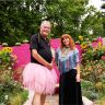 Two people stand in a colorful garden, showcasing the many benefits of gardening. The person on the left wears a pink tutu, while their companion sports a colorful top amidst vibrant foliage and blooming flowers.