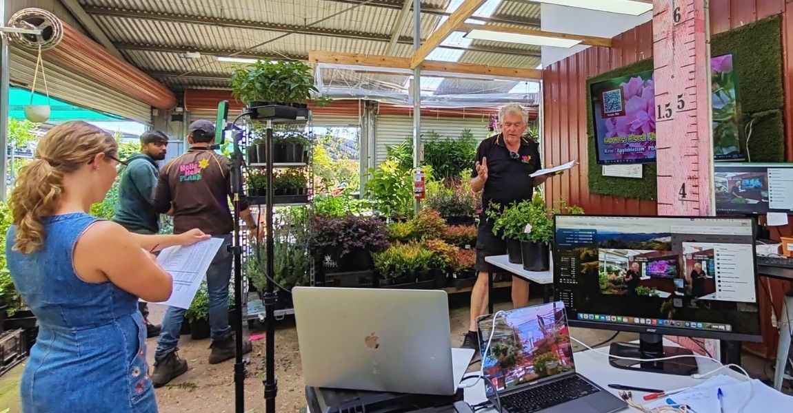 A group of people work in a garden center with computers, plants, and paperwork; one person speaks while others listen and take notes. A sign nearby reads: Truck Drivers Needed.