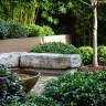 A landscaped garden with dense green shrubs, a tree, a large stone bench, and a shallow bowl water feature in the foreground showcases the beauty of Japanese gardens.