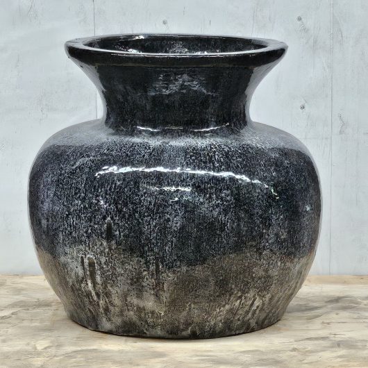 A large, glossy black ceramic vase with a wide body and narrow neck sits on a light wooden surface against a gray background.