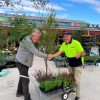 Two men shake hands and smile next to a cart filled with plants in an outdoor garden center. One man gives a thumbs up, showing their confidence that they’ll get the right plants for their garden.