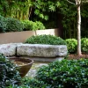 A landscaped garden with dense green shrubs, a tree, a large stone bench, and a shallow bowl water feature in the foreground showcases the beauty of Japanese gardens.